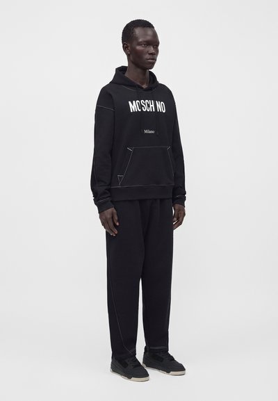 Άνδρας που στέκεται, φοράει μαύρο φούτερ Moschino και μαύρο παντελόνι που ταιριάζει με μαύρα αθλητικά παπούτσια σε ουδέτερο φόντο.
