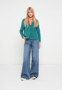 Turkis blazer med to frontlommer, båret over en rød- og hvidternede skjorte. Udsvingede blå jeans og hvide sneakers fuldender looket.