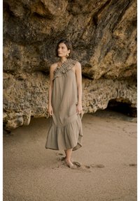 Khaki, ühe õlaga kleit volangidega, voolav kangas ja kihiline ääreosa. Kantud paljajalu rohkel rannaliival, karmi kaljuse taustal.