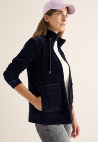 Chaqueta de pana azul marino con un cuello alto, bolsillos frontales y cremallera oculta, combinada con una blusa de color claro y una gorra en tonos pastel.