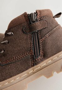 Brūni zamšādi potītes zābaki ar rāvējslēdzēju, rakstainām brūnām auklām un teksturētu gumijas zoli ar zobveida gripu.