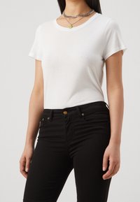 Valkoinen puuvillainen t-paita pyöreällä pääntiellä, lyhyillä hihoilla ja normaalilla istuvuudella, yhdistettynä mustiin kapeisiin farkkuihin, joissa on kultasävyiset metalliosat.