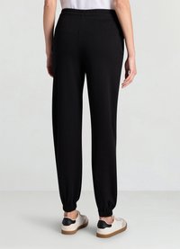 Pantalon de jogging noir avec taille élastique, chevilles resserrées et texture de tissu lisse, associé à des baskets crème et noires.