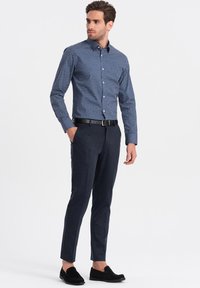 Camicia slim-fit, con bottoni, di colore blu navy con pois bianchi; abbinata a pantaloni scuri e mocassini neri. La texture è liscia e su misura.