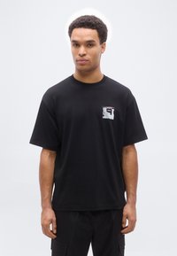 Ανδρας που στέκεται με το πρόσωπο προς τα εμπρός, φοράει μαύρο υπερμεγέθες T-shirt με ένα μικρό αφηρημένο εμπριμέ μπατς και λογότυπο "BOSS" στο στήθος.