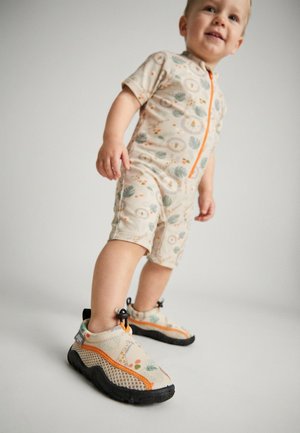 Toddler wearing beige romper and matching sneakers with orange accents and nature-themed patterns, standing against a plain background.