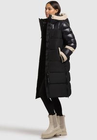 Fekete, hosszú, elegáns kivitelű télikabát, szőrmebélésű kapucnival és kontrasztos bézs mandzsetta díszítéssel. A modell fekete leggingset és bézs csizmát visel.