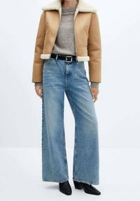 Veste courte en cuir fauve avec col en shearling, pull gris en dessous, jean large bleu clair, ceinture noire avec boucle carrée, et bottines noires.