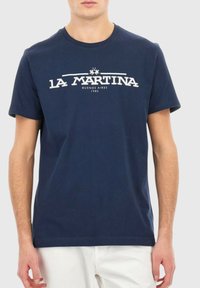 Man i en marinblå La Martina t-shirt med vit logotext över bröstet, i kombination med vita byxor, mot en enfärgad ljus bakgrund.