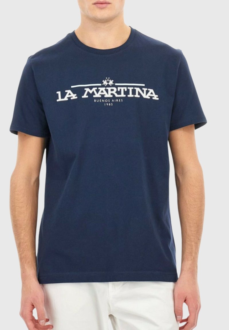 Man i en marinblå La Martina t-shirt med vit logotext över bröstet, i kombination med vita byxor, mot en enf�ärgad ljus bakgrund.