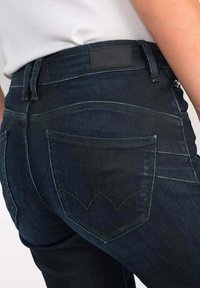 Jean en denim foncé avec une coupe slim, présentant un design de poches arrière texturé et une étiquette noire à la taille. Coutures accentuées visibles.