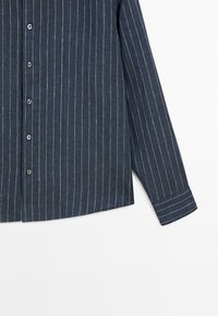 Marineblauw overhemd met witte verticale krijtstrepen, voorkant en rechtermouw met manchetdetail zichtbaar op een witte achtergrond.