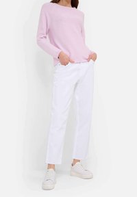 Kvinna som bär en ljus rosa långärmad tröja, vita jeans med rå kant och vita platåskor, stående med händerna i fickorna.