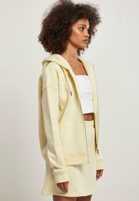 Sudadera con cremallera de color amarillo claro, con mangas largas y bolsillos laterales, combinada con una falda a juego. Tela de algodón, corte relajado, textura suave.