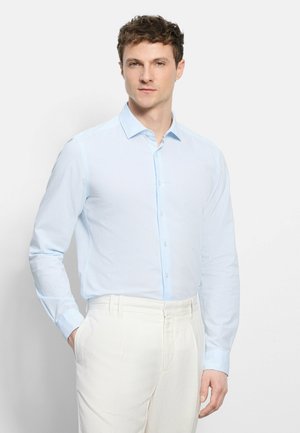 Man wearing light blue long-sleeve button-up shirt tucked into white pants, standing with one hand in pocket against white background.
