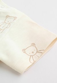 Manche de vêtement bébé couleur crème avec des contours d'ours en peluche marron clair imprimés près du poignet sur un tissu doux.