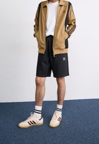 Hnědá atletická bunda s černými pruhy, bílý tričko, černé šortky a krémové tenisky s vínovými akcenty a pruhovanými ponožkami.