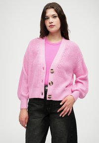 Rosa strikket cardigan med ribbede mansjetter og kant, med fem store dekorative knapper og åpen front. Båret over en rosa skjorte.