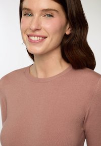 Jasnoróżowy sweter typu crewneck wykonany z miękkiej dzianiny, z prążkowanym dekoltem. Modelka prezentuje naturalny uśmiech i ma falujące ciemne włosy.