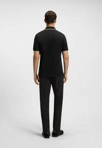 Schwarzes Polohemd mit kurzen Ärmeln, ausgestattet mit einem kontrastierenden weißen Kragenrand, kombiniert mit schwarzen Hosen und schwarzen Schuhen, von hinten betrachtet.