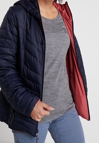 Navy gesteppte Puffjacke mit Kapuze, ausgestattet mit einem roten Futter, Reißverschluss und elastischen Bündchen; getragen über einem grauen Kurzarmshirt.