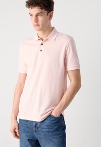 Ροζ πόλο μπλούζα με απαλό υφή, που διαθέτει γιακά, τριπλό κουμπί και μικρό λευκό λογότυπο στο στήθος, συνδυασμένη με μπλε τζιν.