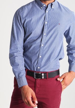 Mann trägt ein blau-weiß gestreiftes Hemd mit Knöpfen, in rote Hose mit schwarzem Gürtel gesteckt, eine Hand in der Tasche.