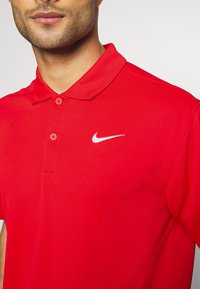Rød polo shirt lavet af glat stof, med krave og to knapper. Et lille hvidt Nike-logo vises på venstre brystområde.