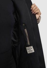 Zwarte buitenschaaljas, met een gladde binnenvoering. Voorzien van een ritszak en een label met de tekst: "Een warme omhelzing gemaakt van stof."