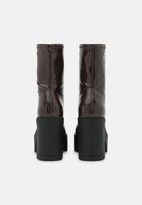 Paire de bottes mi-mollet marron foncé brillant avec des talons carrés épais noirs, vues de dos sur un fond blanc uni.