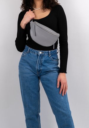 Mujer vestida con camiseta negra de manga larga y vaqueros azules sostiene un bolso gris de bandolera cruzado sobre el pecho frente a un fondo liso.