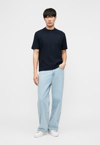 Άνδρας που στέκεται με το ένα χέρι στην τσέπη, φορώντας μαύρο υφασμάτινο κοντομάνικο πουκάμισο, ανοιχτό μπλε τζιν φαρδιάς γραμμής και λευκά αθλητικά παπούτσια.