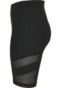 Shorts noirs ajustés taille haute avec deux panneaux en mesh transparent sur la cuisse, vus de profil sur fond blanc.