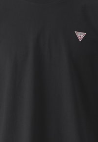 Črna bombažna majica s kratkimi rokavi, ki ima majhen bel in rdeč trikotni logo na levi strani prsi. Kroglično izrezana, gladke teksture in standardne prileganje.