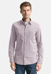 Junger Mann, der ein helllila gestreiftes Hemd über einem weißen Unterhemd und einer blauen Jeans trägt, steht vor einem schlichten Hintergrund.