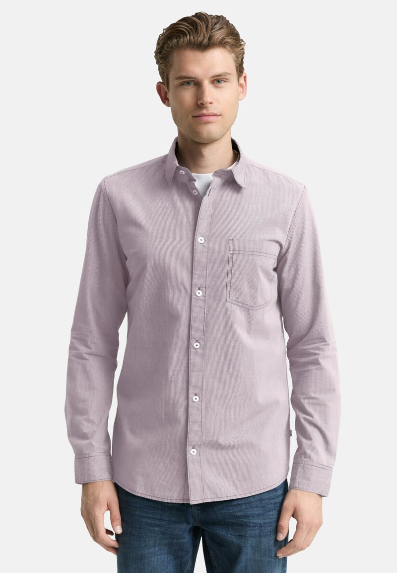 Junger Mann, der ein helllila gestreiftes Hemd über einem weißen Unterhemd und einer blauen Jeans trägt, steht vor einem schlichten Hintergrund.