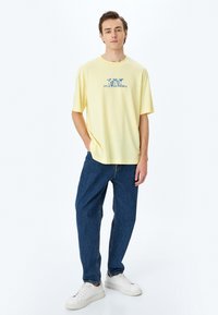 Camiseta de algodón amarilla con un diseño impreso en azul que dice "Fiestas Delicious". Combinada con jeans de mezclilla azules y zapatillas blancas.