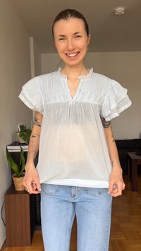 Χαμογελαστή γυναίκα που φοράει μια διάφανη λευκή μπλούζα με βολάν στα μανίκια και ανοιχτό μπλε τζιν, στέκεται σε εσωτερικό χώρο κοντά σε φυτά και έπιπλα.