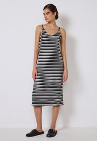 Schwarz-weiß gestreiftes Strickkleid mit dünnen Trägern, figurbetontem Design, knielang, kombiniert mit schwarzen Rutsch-Sandalen. Glatte Stofftextur.