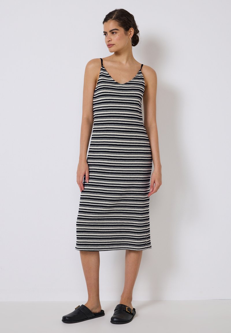 Schwarz-weiß gestreiftes Strickkleid mit dünnen Trägern, figurbetontem Design, knielang, kombiniert mit schwarzen Rutsch-Sandalen. Glatte Stofftextur.