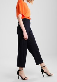 Pomarańczowa bluzka z krótkim rękawem w połączeniu z granatowymi, dopasowanymi spodniami z wysokim stanem. Stylizację dopełniają czarne sandały na wysokim obcasie z paskami.