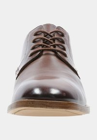 Chaussure habillée en cuir marron avec un bout rond, des lacets plats et une surface lisse et polie. Semelle marron clair avec une légère texture.