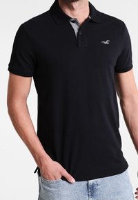 Černé polo tričko s krátkými rukávy, se šedým límcem a vyšitým logem na hrudi. Vyrobeno z hladkého materiálu s klasickým pohodlným střihem.