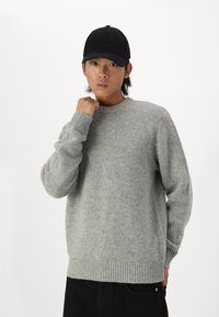 Gai pelēks adīts džemperis ar ribinātu apkakli un aprocēm, piedāvājot maigu tekstūru. Valkāts ar melnu cepuri un tumšajiem biksēm.