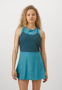 Frau mit lockigem blondem Haar, die ein ärmelloses teal-farbenes Sportkleid mit Nike-Logo trägt, vor einem schlichten weißen Hintergrund stehend.