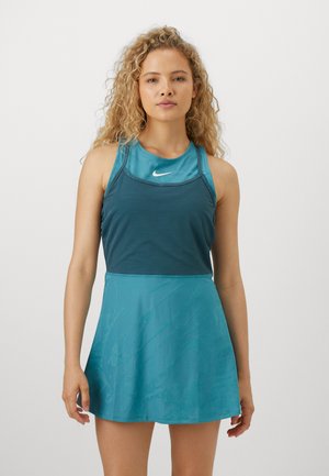 Frau mit lockigem blondem Haar, die ein ärmelloses teal-farbenes Sportkleid mit Nike-Logo trägt, vor einem schlichten weißen Hintergrund stehend.