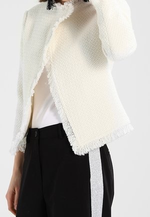 Femme portant une veste écrue texturée à franges sur un top blanc et un pantalon noir avec une bande latérale argentée pailletée.
