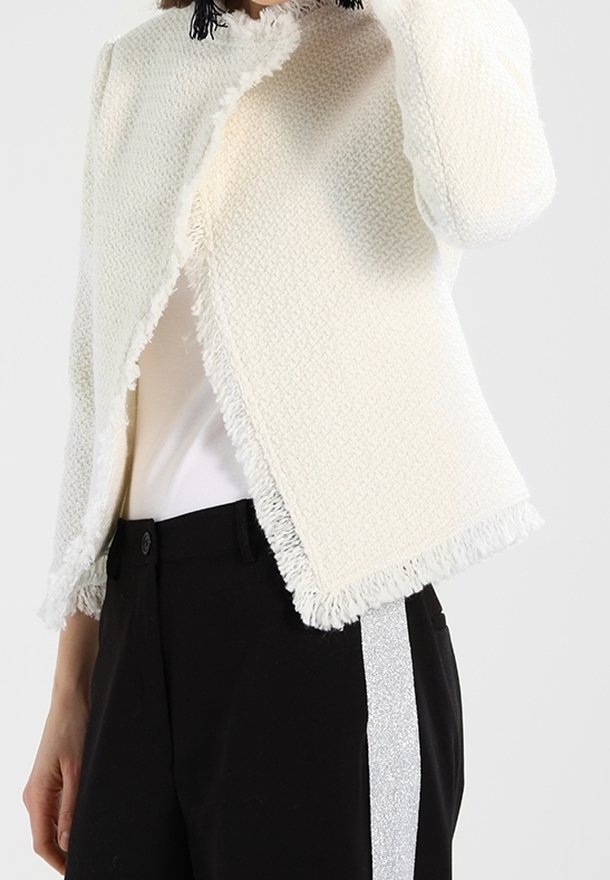 Femme portant une veste écrue texturée à franges sur un top blanc et un pantalon noir avec une bande latérale argentée pailletée.