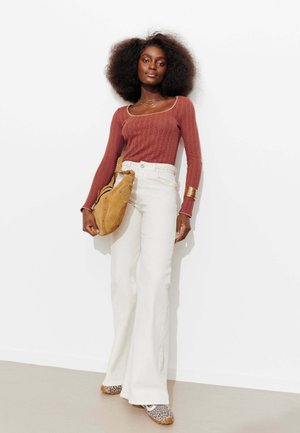 Femme avec une coiffure afro portant un haut à manches longues couleur rouille, un pantalon large taille haute blanc, des chaussures imprimé léopard, et tenant un sac en daim couleur beige.