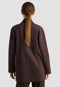 Frau mit langen braunen Haaren, die zu einem tiefen Pferdeschwanz gebunden sind, trägt ein dunkelbraunes maßgeschneidertes Blazer und passende Hose, steht vor einem schlichten Hintergrund.